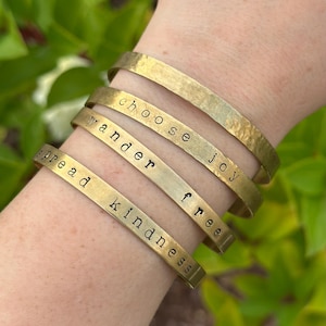 May include: A collection of gold-toned metal bracelets, each engraved with a different inspirational word: "spread kindness", "wander", "free", and "choose joy". The bracelets are stacked on a wrist, showcasing their simple, elegant design.