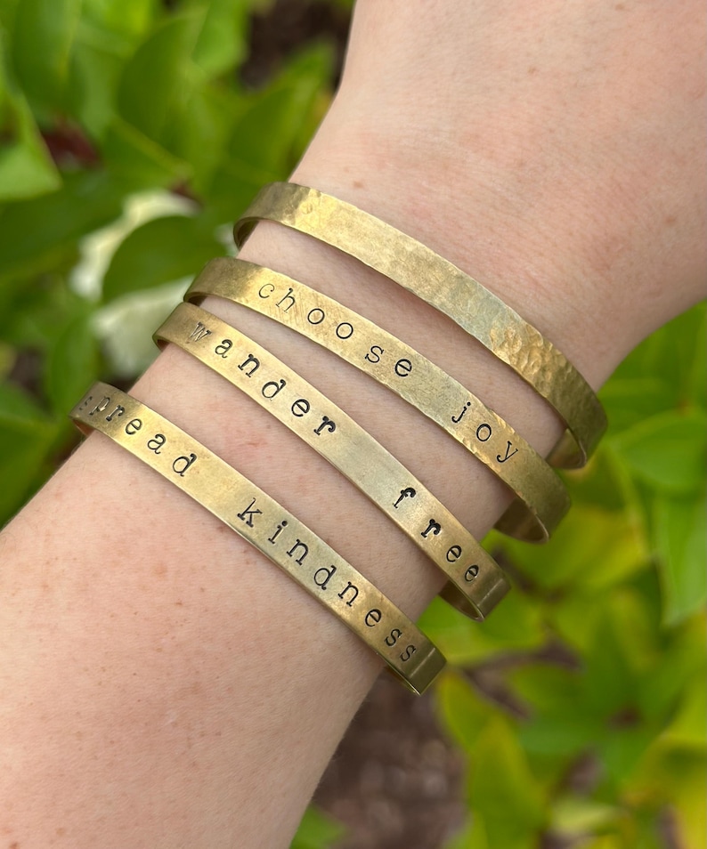 May include: A collection of gold-toned metal bracelets, each engraved with a different inspirational word: "spread kindness", "wander", "free", and "choose joy". The bracelets are stacked on a wrist, showcasing their simple, elegant design.