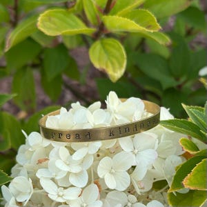 May include: A gold-colored metal bracelet with the words "spread kindness" stamped on it. The bracelet rests on a bed of white flowers, with green foliage in the background.
