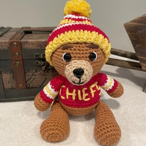 May include: A crocheted brown bear wearing a red and yellow striped hat with a yellow pom-pom and a red sweater with white stripes and the word "CHIEF" in yellow lettering.