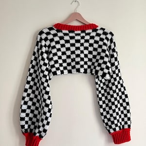 Może przedstawiać: Szydełkowy bolerko w czarno-białą szachownicę z długimi rękawami. Sweter ma czerwony kołnierz i mankiety. Wisi na różowym wieszaku na białym tle.