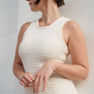 Op de afbeelding: Een vrouw die een witte, textuurrijke, mouwloze jurk draagt met een gouden ketting en oorbellen. Haar haar is naar achteren getrokken in een knot en ze draagt een gouden haarspeld. Ze staat voor een witte muur.