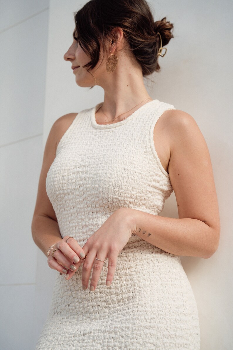 Op de afbeelding: Een vrouw die een witte, textuurrijke, mouwloze jurk draagt met een gouden ketting en oorbellen. Haar haar is naar achteren getrokken in een knot en ze draagt een gouden haarspeld. Ze staat voor een witte muur.