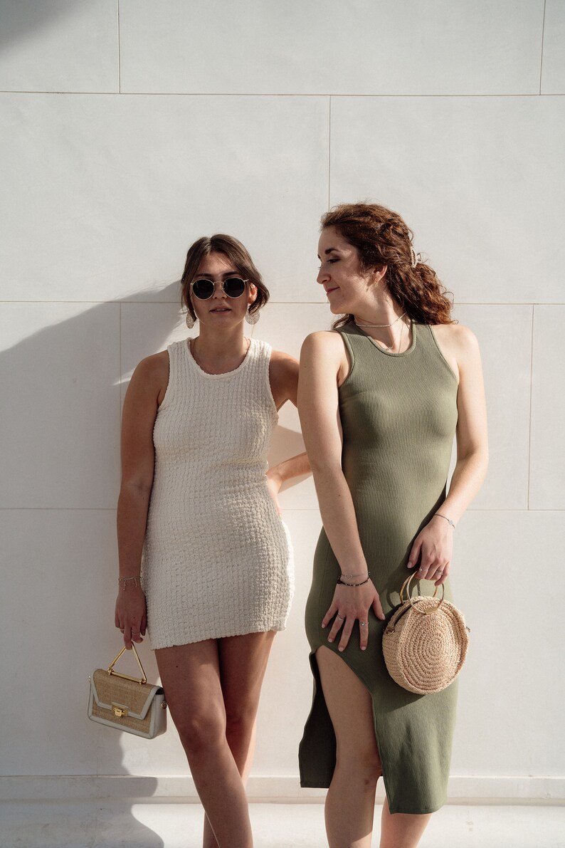 Op de afbeelding: Twee mensen in zomerjurken staan voor een witte betegelde muur. De persoon links draagt een witte jurk en een zonnebril. De persoon rechts draagt een groene jurk en heeft een rieten tas.