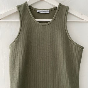 Op de afbeelding: Een groene geribbelde tanktop met een wit label met de tekst "#SPAWNYOURSELF" op een witte hanger.