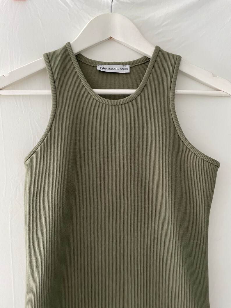 Op de afbeelding: Een groene geribbelde tanktop met een wit label met de tekst "#SPAWNYOURSELF" op een witte hanger.
