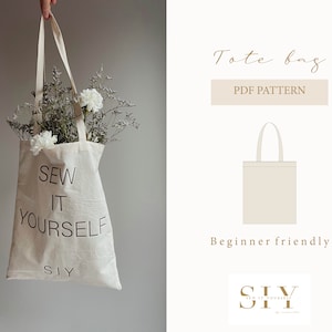 May include: A white tote bag with the text "Sew It Yourself" and the abbreviation "SIY" printed on it. The bag is being held by a hand and has dried flowers inside.