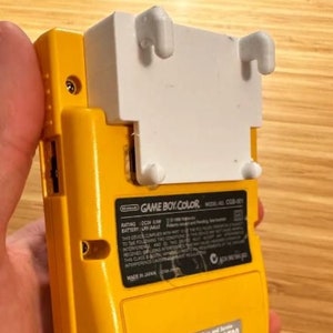 Puede incluir: Una consola de videojuegos portátil Game Boy Color amarilla con una placa frontal blanca. La consola tiene una pantalla pequeña, botones y una ranura para cartuchos. El texto "GAME BOY COLOR" está impreso en la parte frontal de la consola.