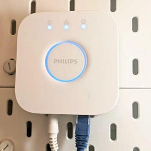 Op de afbeelding: Een witte Philips smart home hub met een blauwe licht ring rond de middelste knop. De hub heeft drie blauwe indicator lampjes aan de bovenkant. De hub is gemonteerd op een witte pegboard muur met twee kabels die in de onderkant zijn gestoken.