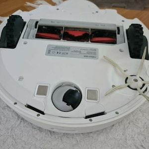 Op de afbeelding: Witte robotstofzuiger met een zwarte borstelrol en twee zijborstels. De stofzuiger heeft een stofreservoir en een zijborstelbevestiging. De stofzuiger staat op een wit tapijt.