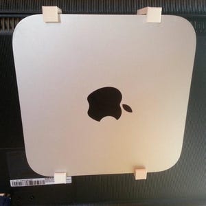 May include: A silver Mac mini computer with the Apple logo on the front. The computer is mounted on the back of a black television screen with white plastic clips.
