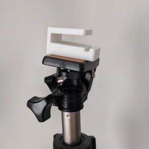May include: A white, U-shaped camera accessory is mounted on a black tripod head. The tripod has a silver pole and black adjustment knobs. The accessory appears to be made of plastic and is designed to hold a camera or phone.