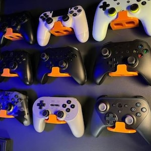 Puede incluir: Un expositor de pared de ocho mandos de videojuegos diferentes. Los mandos son de diferentes colores y estilos, incluyendo negro, blanco, gris y camuflaje. Los mandos están montados en soportes de plástico naranja.