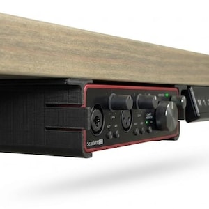 Könnte beinhalten: Eine schwarze 3D-gedruckte Untertischhalterung für ein Focusrite Scarlett Audio-Interface mit roter Frontblende, mehreren Drehreglern und Eingangsanschlüssen. Die Halterung ist an der Unterseite einer hölzernen Tischplatte neben einem Tastenfeld befestigt.