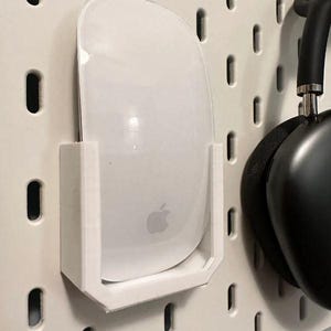 Könnte beinhalten: Eine weiße kabellose Maus mit einem Apple-Logo ist an einer weißen Lochwand montiert. Die Maus befindet sich in einem weißen Kunststoffhalter.