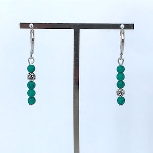 Può includere: Un paio di orecchini pendenti con perline color turchese e dettagli argentati. Ogni orecchino presenta una serie di perline turchesi, una perlina decorativa argentata e una chiusura a leva. Sfondo bianco.