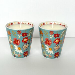 Puede incluir: Dos velas de color turquesa en tazas de cerámica con estampado floral. Las tazas presentan un patrón repetido de flores rojas, blancas y amarillas con hojas verdes. Cada vela tiene cera blanca y una sola mecha.