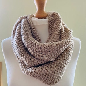 May include: A beige, chunky knit infinity scarf on a mannequin. The scarf is made of thick yarn, creating a textured appearance. The neutral colour makes it a versatile accessory. The scarf is designed to be worn looped around the neck, a popular winter accessory.