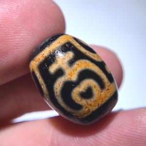 Puede incluir: Primer plano de una cuenta ovalada con un diseño en espiral negro y marrón. La cuenta se sostiene entre dos dedos. La cuenta tiene un acabado brillante y un diseño único.