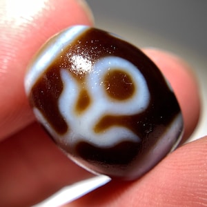 Puede incluir: Primer plano de una cuenta dzi marrón y blanca. La cuenta tiene un fondo marrón oscuro con un patrón blanco. La cuenta es de forma ovalada y parece lisa. El patrón blanco es un diseño estilizado.