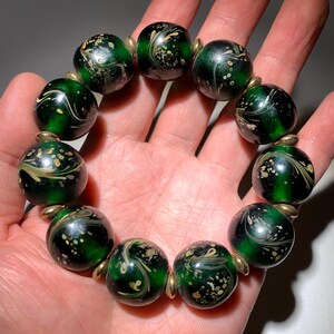 Puede incluir: Un brazalete de vidrio verde con motas y remolinos dorados. El brazalete está hecho de cuentas redondas y tiene un cierre dorado.