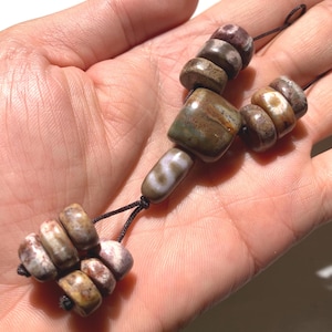 Puede incluir: Un collar de cuentas con cuentas de piedra natural en tonos marrón, beige y crema. Las cuentas están ensartadas en un cordón negro, con una mezcla de formas redondeadas y cuadradas. El collar se muestra en una mano.