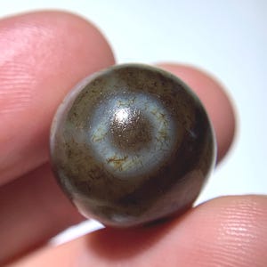 Puede incluir: Una piedra esférica de color marrón oscuro con una superficie pulida. La piedra tiene un patrón único de marcas claras en forma de telaraña. La esfera se sostiene entre dos dedos, mostrando su tamaño y textura. La piedra parece ser un objeto decorativo.