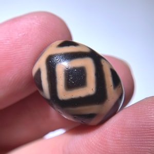Puede incluir: Primer plano de una cuenta Dzi, un tipo de cuenta de ágata tibetana. La cuenta de forma ovalada presenta un diseño geométrico negro y beige. La cuenta se sostiene entre dos dedos.
