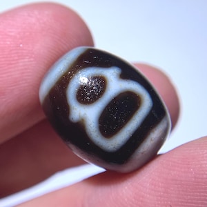 Puede incluir: Primer plano de una cuenta dzi marrón y blanca. La cuenta de forma ovalada presenta un fondo marrón oscuro con un patrón blanco en forma de ojo. La cuenta se sostiene entre dos dedos.