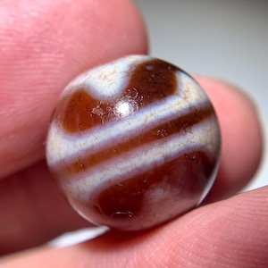 Puede incluir: Una cuenta redonda de ágata marrón con rayas blancas. La cuenta es lisa y pulida.