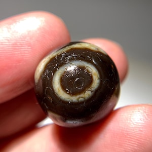 Puede incluir: Una cuenta de ágata marrón oscuro y blanco con un patrón de remolino. La cuenta tiene una superficie lisa y pulida.