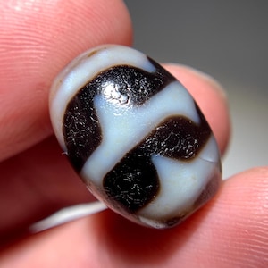 Puede incluir: Una cuenta de ágata blanca y negra con un patrón de remolino. La cuenta es de forma ovalada y tiene una superficie lisa y pulida.
