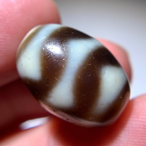 Puede incluir: Una cuenta de ágata blanca y marrón con un patrón de remolino. La cuenta es lisa y pulida.