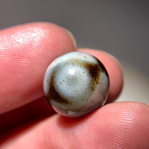 Puede incluir: Una cuenta de ágata blanca y marrón con un patrón de remolino. La cuenta es redonda y lisa.