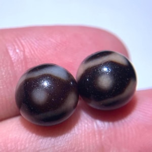 Puede incluir: Dos cuentas redondas y pulidas con una base de color marrón oscuro y patrones blancos en forma de ojo. Las cuentas son lisas y reflectantes, con un acabado brillante. El fondo es borroso y de color claro.