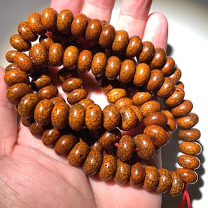 Puede incluir: Un collar de cuentas de oración. Las cuentas son de color marrón anaranjado, de forma ovalada y con motas de color marrón oscuro. Están ensartadas en un cordón rojo. Las cuentas se sostienen en una mano.