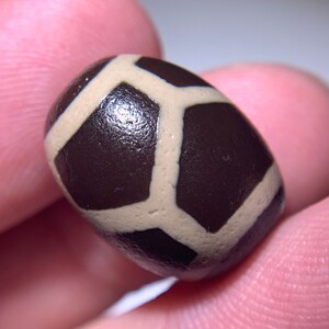 Puede incluir: Una cuenta de ágata marrón y blanca con un patrón geométrico. La cuenta es lisa y pulida.