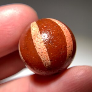 Puede incluir: Una canica redonda de ágata roja con rayas blancas. La canica es lisa y pulida.