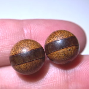 Puede incluir: Dos cuentas redondas marrones con una banda marrón oscuro alrededor del centro. Las cuentas tienen una apariencia texturizada y agrietada. Las cuentas se sostienen en la mano de una persona.