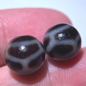 Puede incluir: Dos cuentas redondas, de color marrón oscuro, con un acabado brillante. Cada cuenta presenta un patrón geométrico blanco. Las cuentas se sostienen en la mano de una persona, con la piel rosada de la mano visible en el fondo.