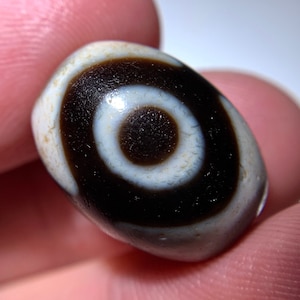 Puede incluir: Una cuenta de ágata blanca y negra con un patrón circular. La cuenta tiene un acabado brillante y es de forma ovalada.