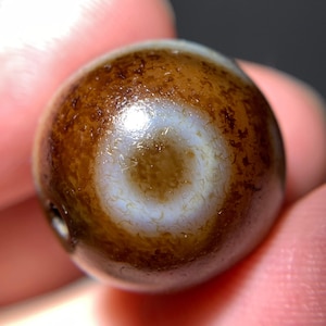 Puede incluir: Una cuenta de ágata marrón con un patrón de ojo blanco. La cuenta tiene un agujero perforado para encordar.