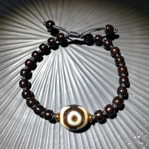 Puede incluir: Un brazalete de cuentas de madera marrón con un dije en forma de ojo blanco y marrón. El dije está rodeado de detalles dorados.