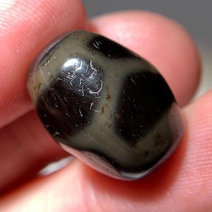 Puede incluir: Una cuenta de ágata negra y gris con intrincados patrones y marcas. La cuenta es lisa y pulida, con un acabado brillante.