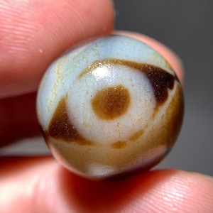Puede incluir: Una cuenta de ágata blanca con marcas marrones que se asemejan a un ojo. La cuenta tiene una superficie lisa y pulida y está ligeramente agrietada.