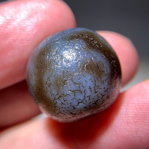 Puede incluir: Una cuenta redonda de ágata de color gris oscuro con un patrón blanco en espiral. La cuenta tiene una superficie lisa y pulida.