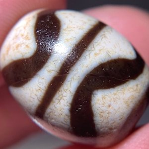 Puede incluir: Una cuenta de ágata blanca y marrón con un patrón de remolino. La cuenta es lisa y pulida.