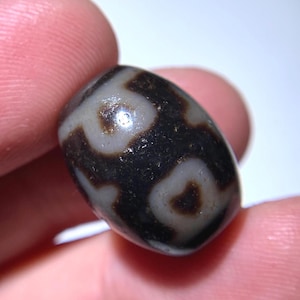 Puede incluir: Primer plano de una cuenta oscura, de forma ovalada, con un patrón de remolinos grises claros y marrones. La cuenta se sostiene entre dos dedos, mostrando su superficie pulida y su intrincado diseño. El fondo oscuro de la cuenta contrasta con los remolinos más claros.