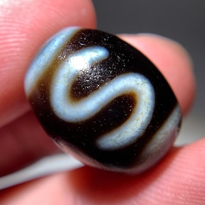 Puede incluir: Una cuenta de ágata negra y blanca con un patrón de remolino. La cuenta es lisa y pulida.