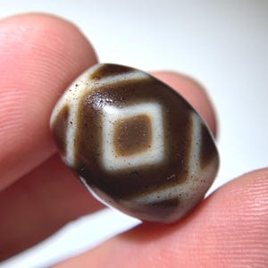 Puede incluir: Primer plano de una cuenta dzi, un tipo de cuenta de ágata. La cuenta ovalada presenta un patrón geométrico marrón y blanco. La cuenta se sostiene entre dos dedos.